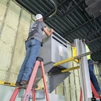 Worker on a red ladder servicing equipment on an industrial ceiling with exposed ductwork